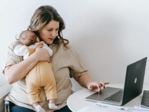 A woman sits in front of a laptop, typing with one hand while holding a newborn with the other.